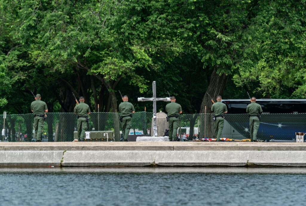 Military personnel participate in a solemn ceremony near a cross monument in Washington D.C. park.